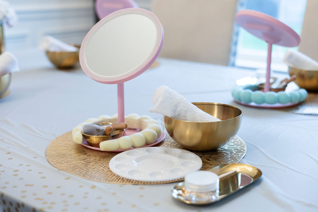Makeup mirror with pink stand on a table with various items including a bowl and a container.