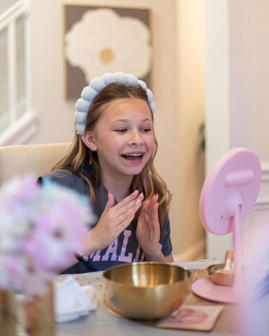 Young girl smiling and applying skincare at a table with a pink lamp and gold bowl.