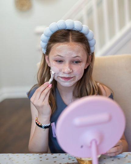 Young girl applying facial mask with a hairband on her head, sitting in front of a mirror.