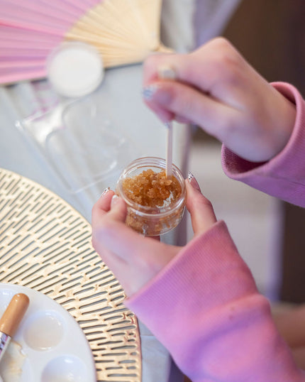 Person holding a small container of brown sugar scrub with a brush over it, on a table.