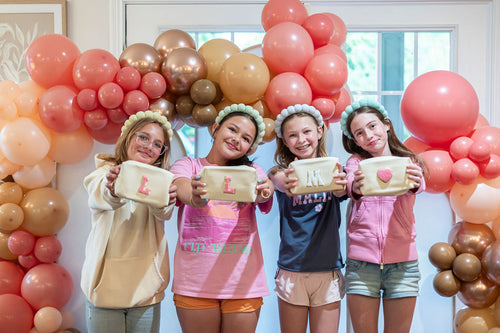 Four children holding makeup bags with initials standing in front of a colorful balloon arch.