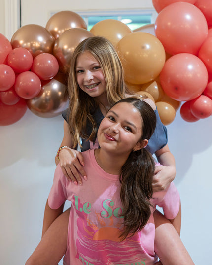 Two young girls posing together with pink, gold, and red balloons in the background.