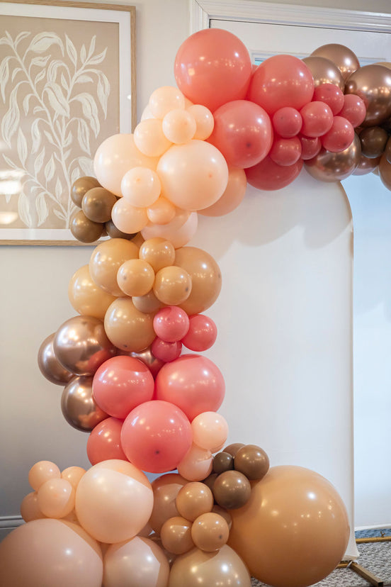 Balloon arch in pink, gold, and white colors against a neutral wall.