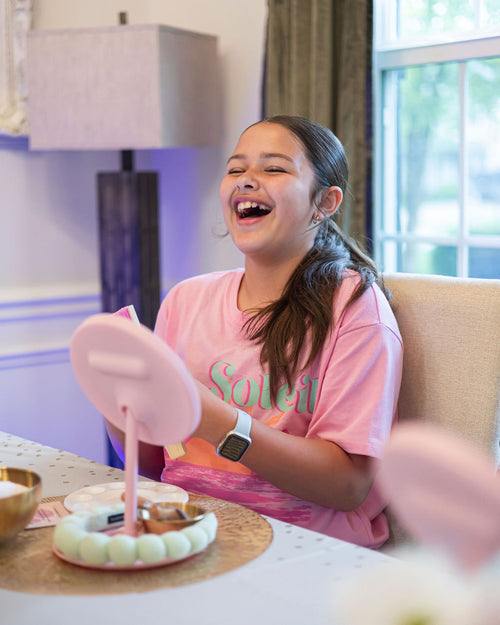 Girl in a pink shirt sitting at a table with a mirror laughing